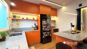Cozinha planejada com armários na cor laranja vibrante, bancada em granito cinza, ilha central com cooktop e iluminação em fita de LED sob os armários aéreos. Projeto Simonetto Grupo Duarte.